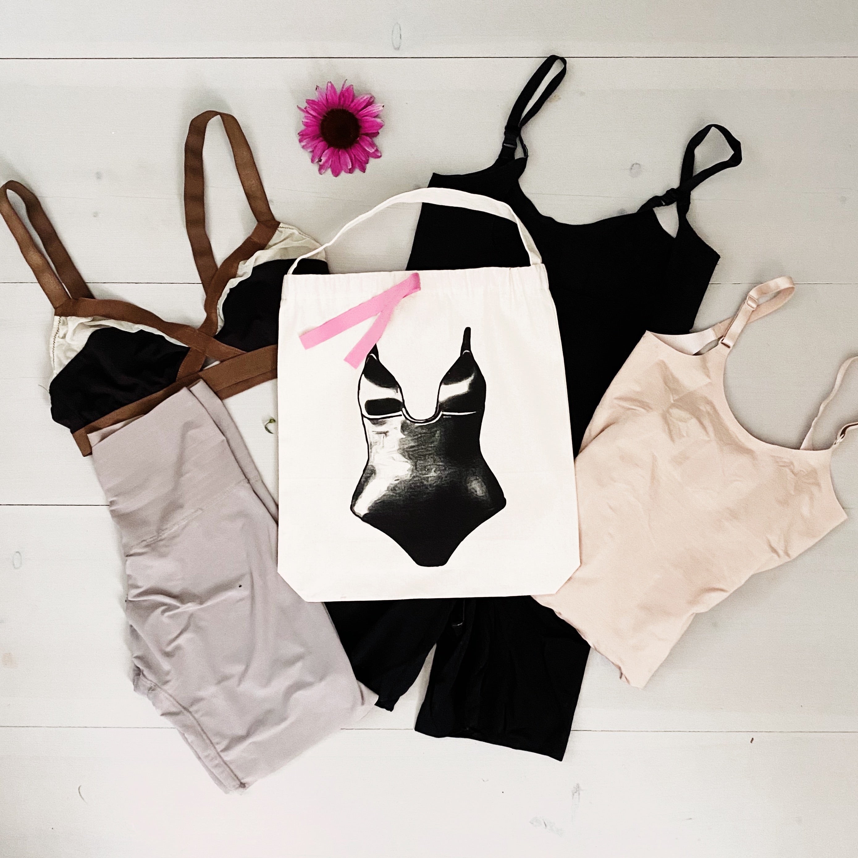 Bag-all Shapewear Travel Bag in cream canvas featuring black swimsuit print with pink bow, displayed with assorted shapewear pieces and pink flower on white wooden background