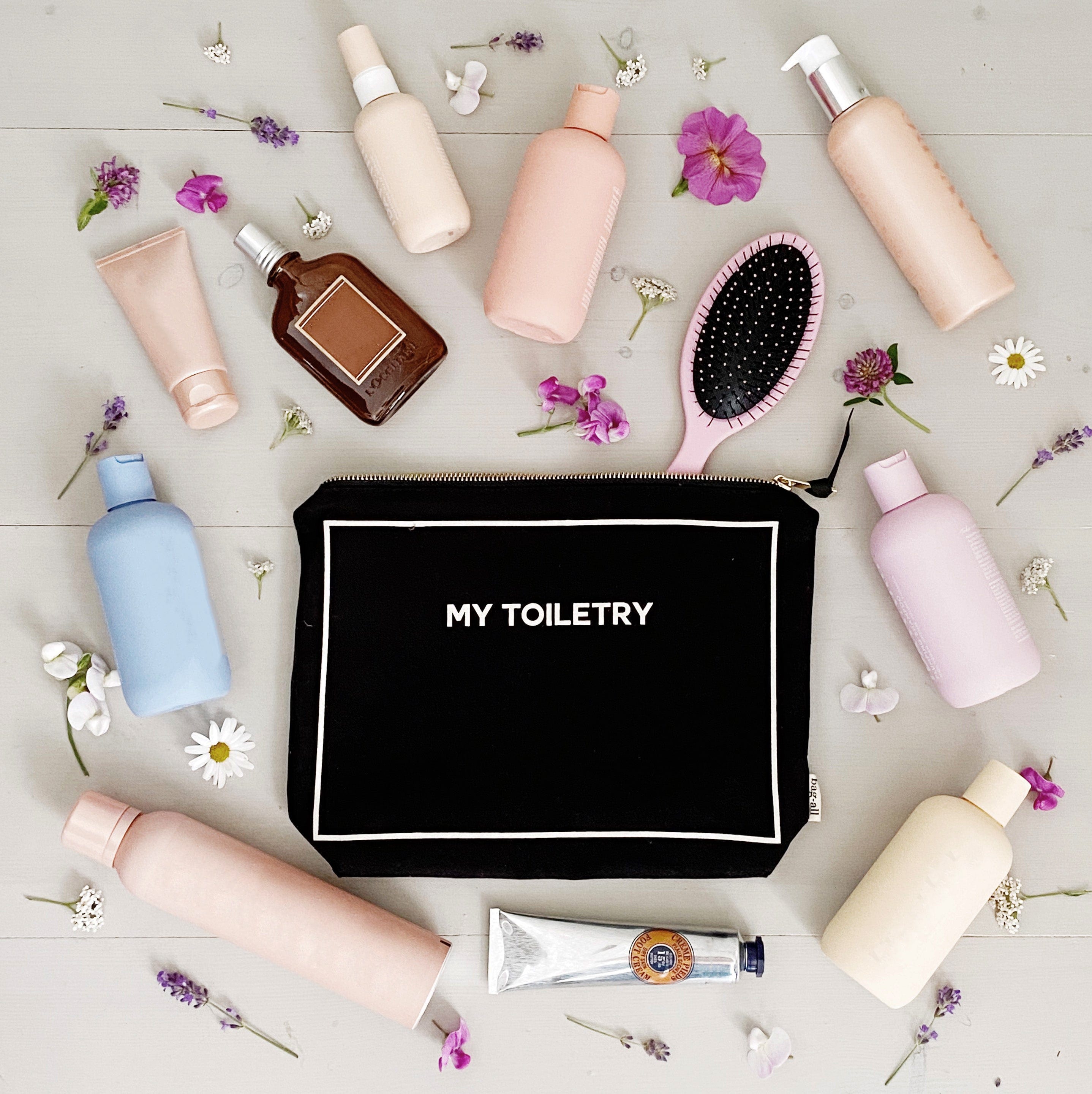 Bag-all Weekend Essential Packing Set features black toiletry pouch surrounded by pastel beauty products, hairbrush, and delicate flowers on light wooden surface