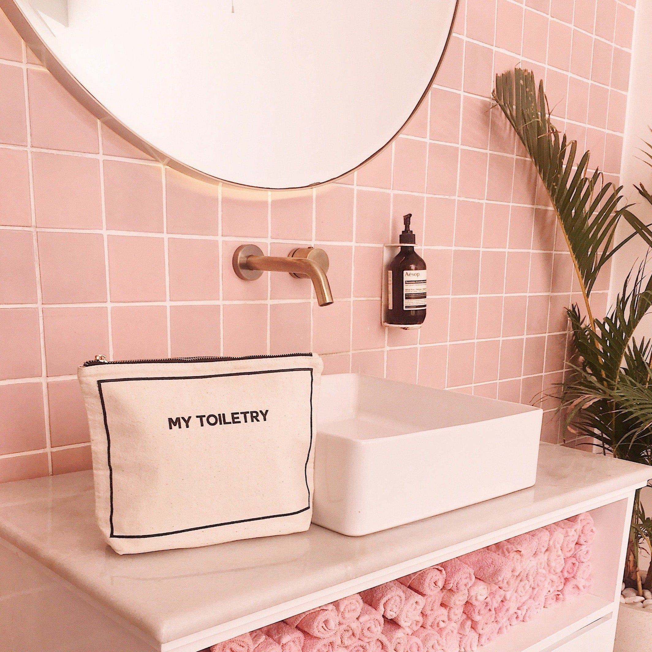 Bag-all Weekend Essential Packing Set in cream featuring organized cotton pouches in modern bathroom setting with pink tiles, gold fixtures and palm decor, showcasing toiletry bag
