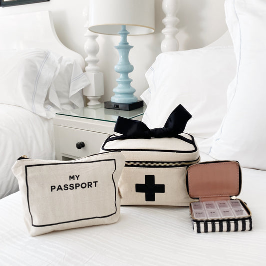 Bag-all First Aid Travel Gift Set 3-Pack in cream: stylish cotton passport pouch, medical storage box with black bow, and striped pill case arranged on white bedding