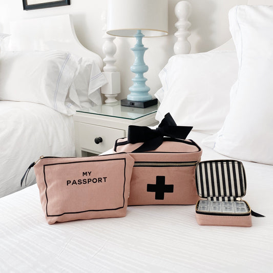 Bag-all First Aid Travel Gift Set 3-Pack in pink featuring passport pouch, medical storage box with black cross, and striped pill case on white bedding, perfect for organized travel