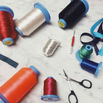 Bag-all monogram customization supplies featuring colorful thread spools, scissors, and ribbons for personalized bag embroidery, showing monogramming tools and materials on marble surface