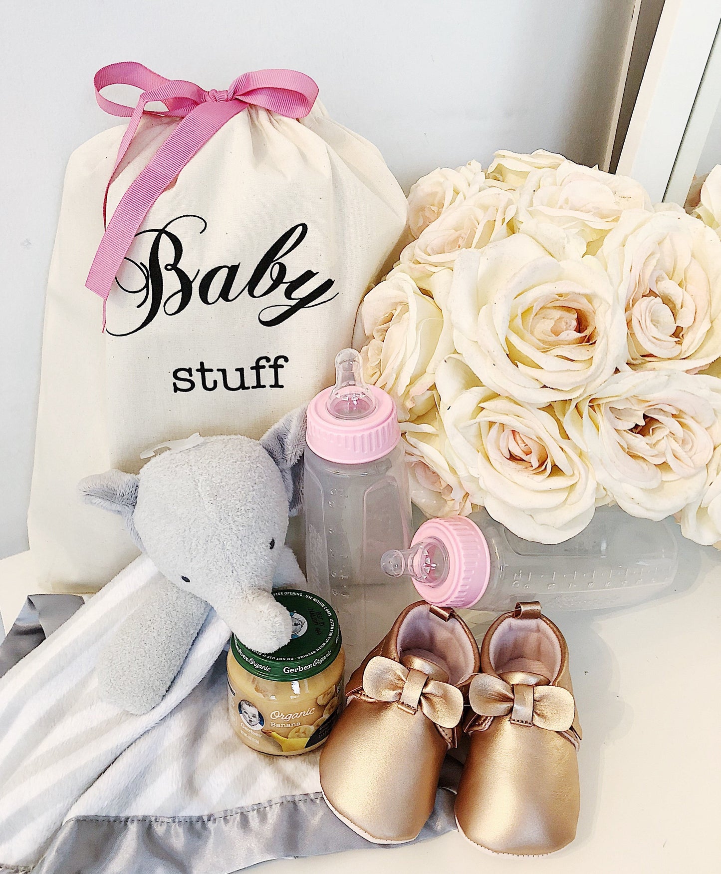 Bag-all Baby Stuff organizer bag with pink bow, displayed with gold baby shoes, plush elephant, baby bottles and cream roses - perfect nursery storage solution