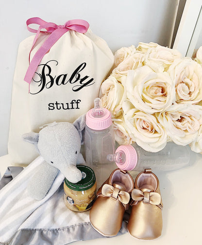 Bag-all Baby Stuff organizer bag with pink bow, displayed with gold baby shoes, plush elephant, baby bottles and cream roses - perfect nursery storage solution