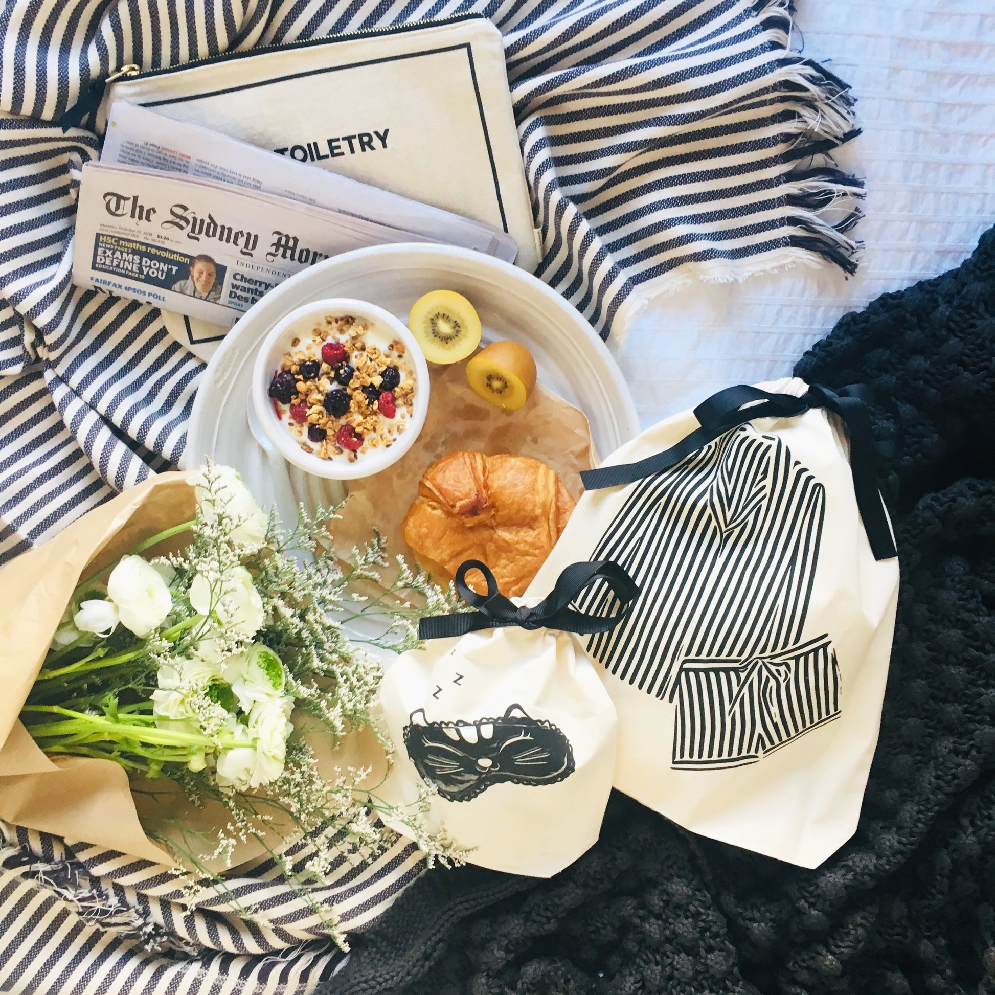 Bag-all black and white striped travel bags with breakfast setting, fresh flowers, croissant, fruits and muesli bowl, Sydney Morning newspaper