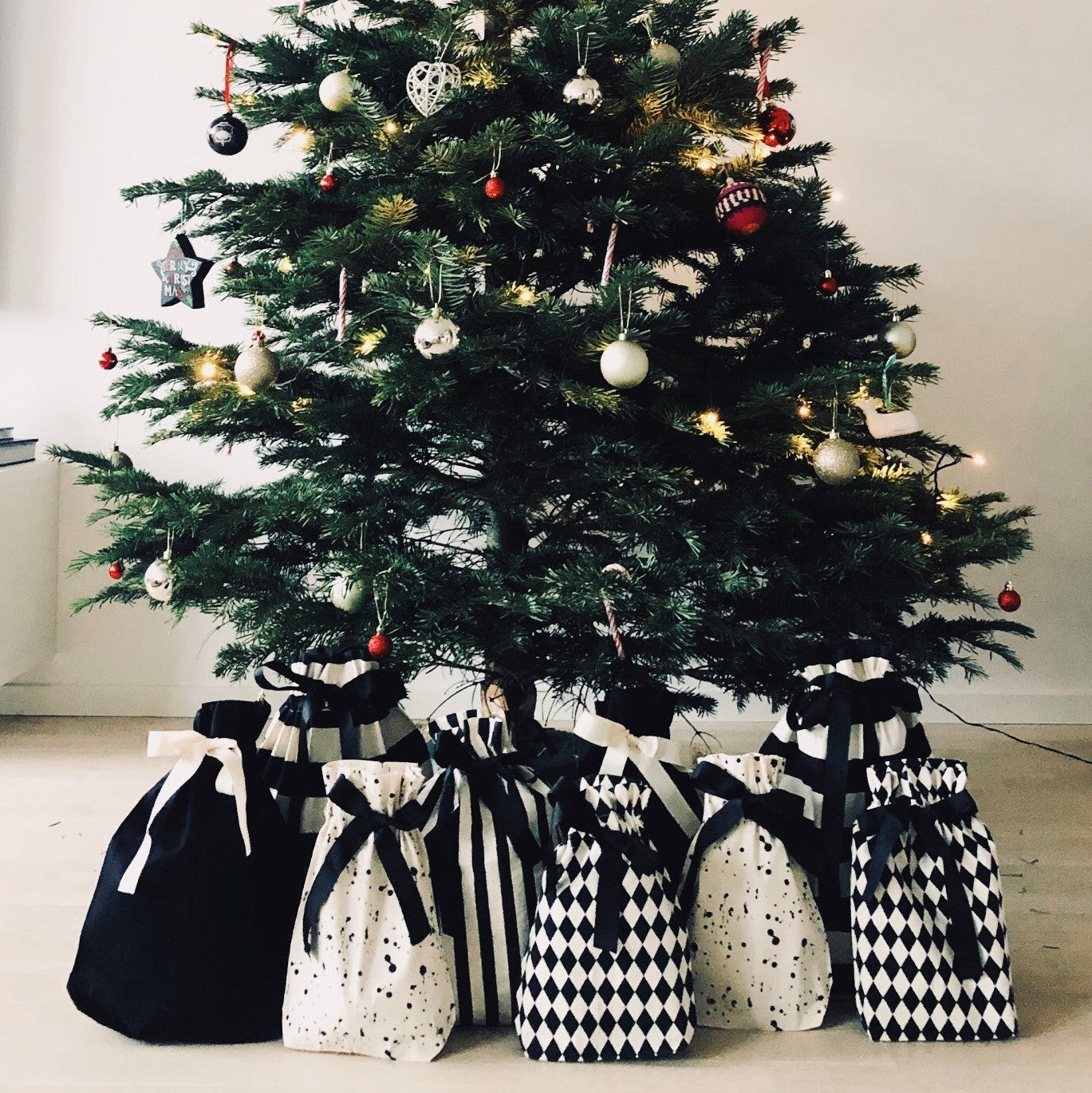 Bag-all Gift Bag Striped Medium shown with other black and white patterned gift bags beneath decorated Christmas tree, showcasing sustainable gift wrapping alternatives for holiday season