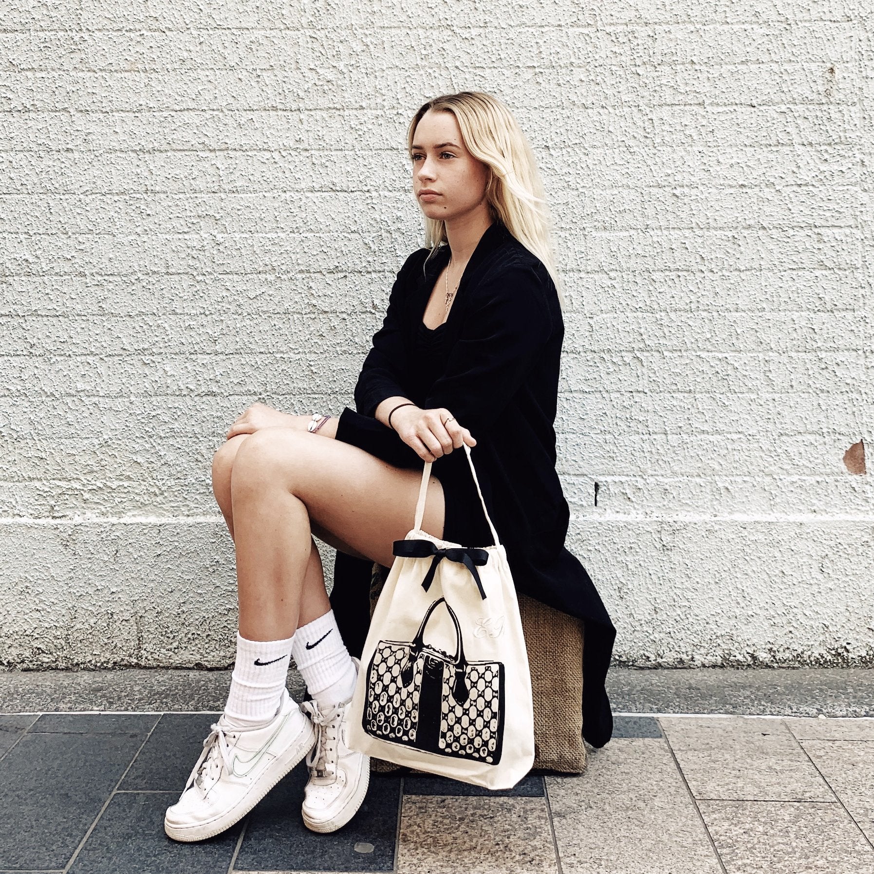 A person seated outdoors displaying Bag-all's Vintage Handbag Travel Bag in Cream with black trim, paired with a black coat and white sneakers against a textured white wall background