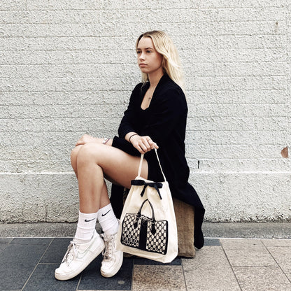 A person seated outdoors displaying Bag-all's Vintage Handbag Travel Bag in Cream with black trim, paired with a black coat and white sneakers against a textured white wall background