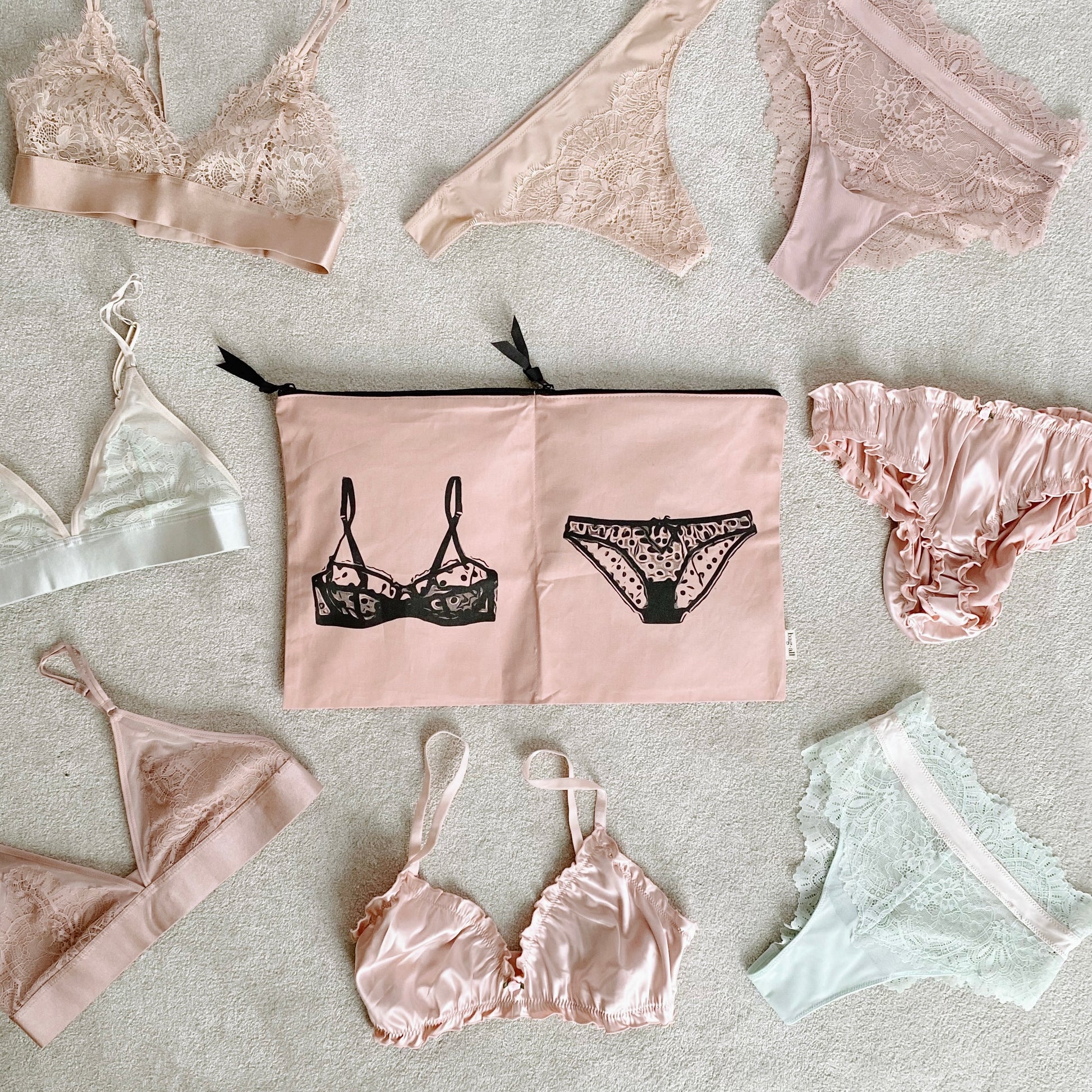 Bag-all Double Lingerie Packing Pouch in pink/blush with black lingerie print, surrounded by delicate lace undergarments in pastel pink and white tones, featuring dual compartments and black zipper closures
