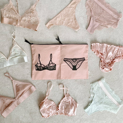 Bag-all Double Lingerie Packing Pouch in pink/blush with black lingerie print, surrounded by delicate lace undergarments in pastel pink and white tones, featuring dual compartments and black zipper closures