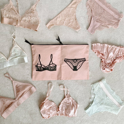 Bag-all Double Lingerie Packing Pouch in pink/blush with black lingerie print, surrounded by delicate lace undergarments in pastel pink and white tones, featuring dual compartments and black zipper closures