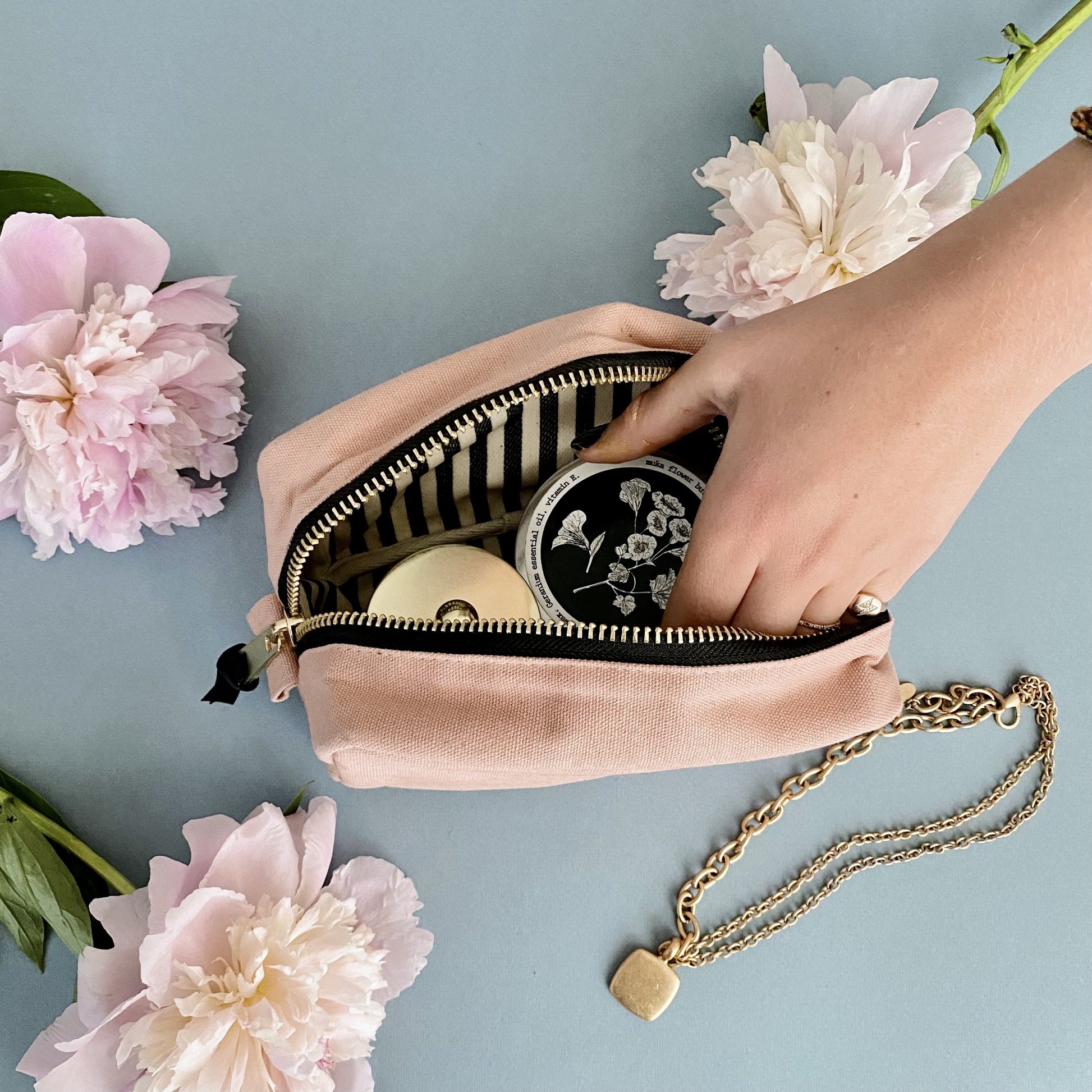 Bag-all Small Toiletry Pouch in Pink/Blush with striped interior lining, gold chain strap, and black zipper, styled with pink peonies against blue background