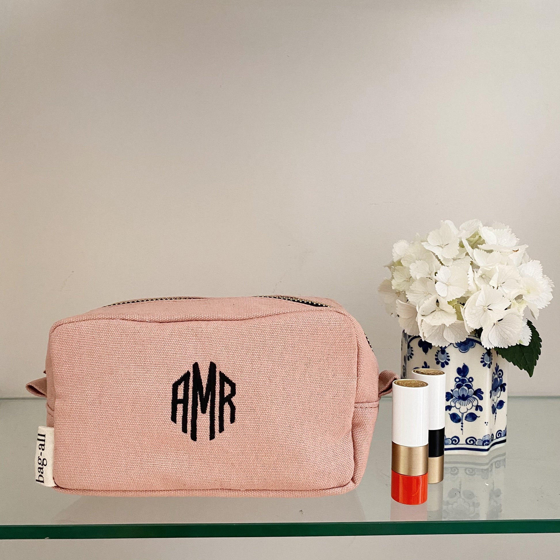 Bag-all customizable pink makeup pouch with monogram AMR, displayed next to white hydrangeas in blue and white vase and lipstick on glass shelf