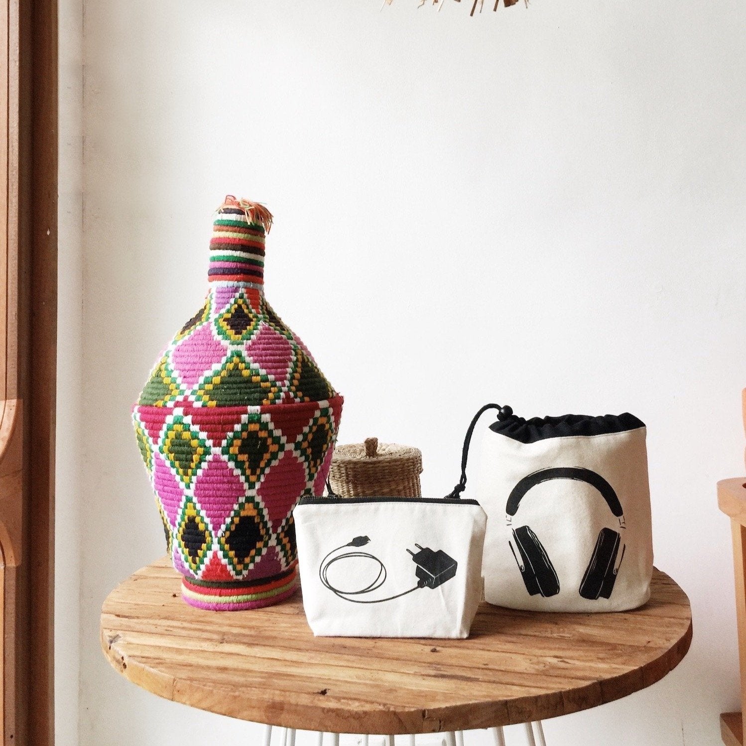 Bag-all Cream Charger Pouch with headphone print design, displayed on wooden table alongside decorative woven bottle, organized storage solution for cables and electronics