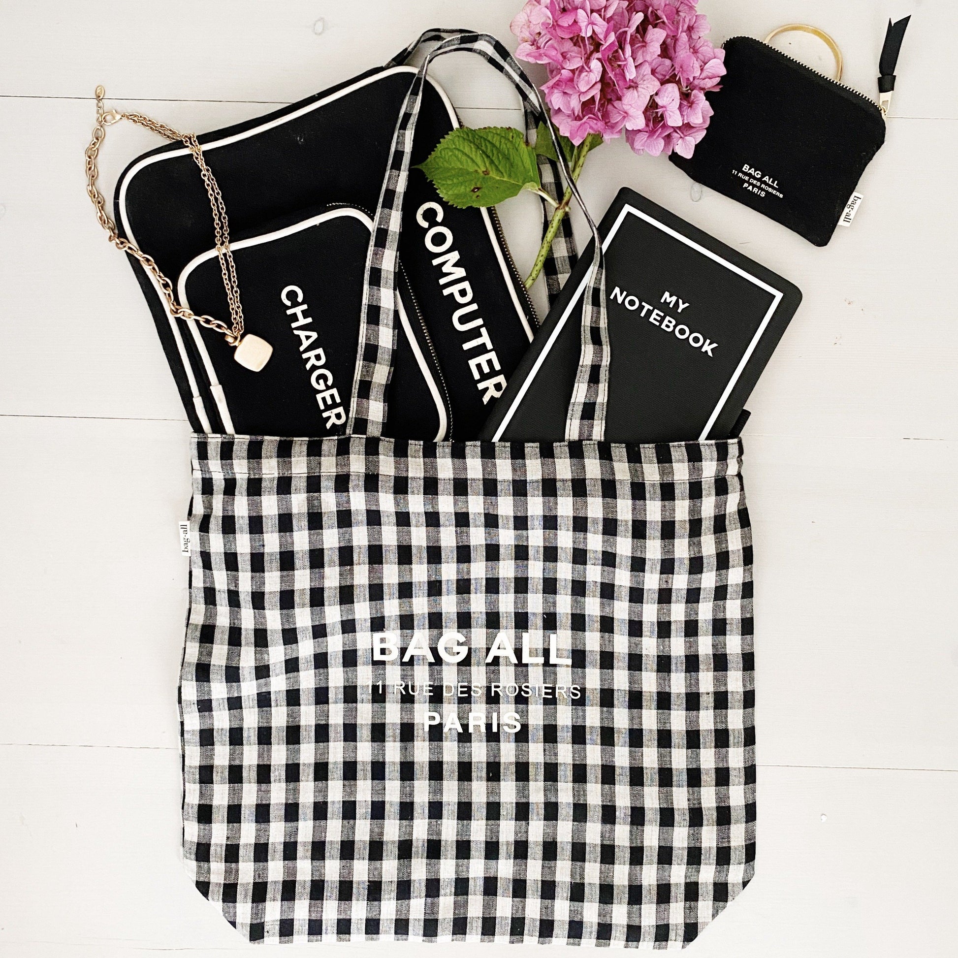Bag-all Paris Le Marais Tote in black and white gingham pattern, filled with essentials including notebook and electronics case, styled with pink hydrangea flower on white wooden surface