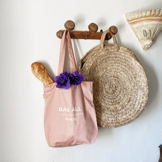 Bag-all Paris Le Marais pink cotton tote bag with zipper closure hanging on wooden hooks, styled with purple flowers, French baguette, and natural straw basket bag for everyday essentials