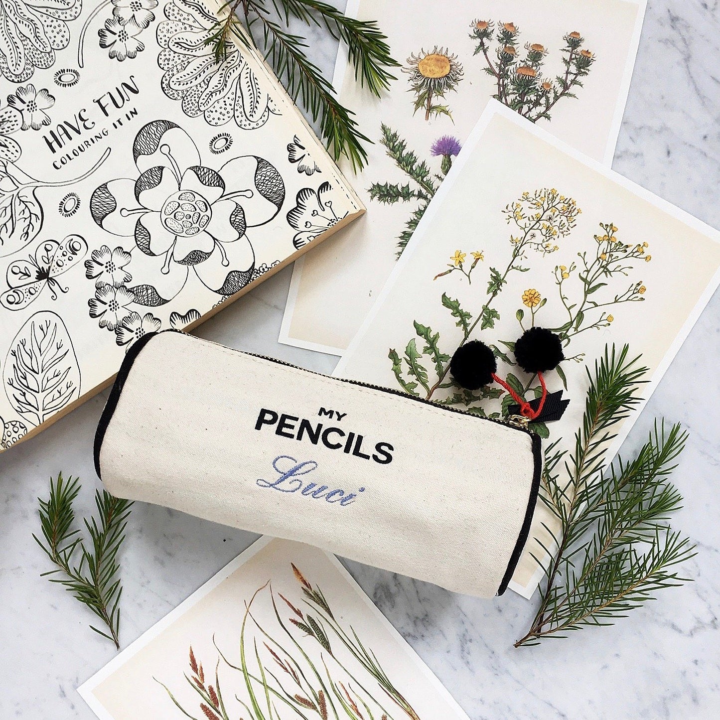 Bag-all cream canvas pencil case with black text reading My Pencils, decorated with pom-poms, displayed on marble surface with botanical illustrations and pine sprigs