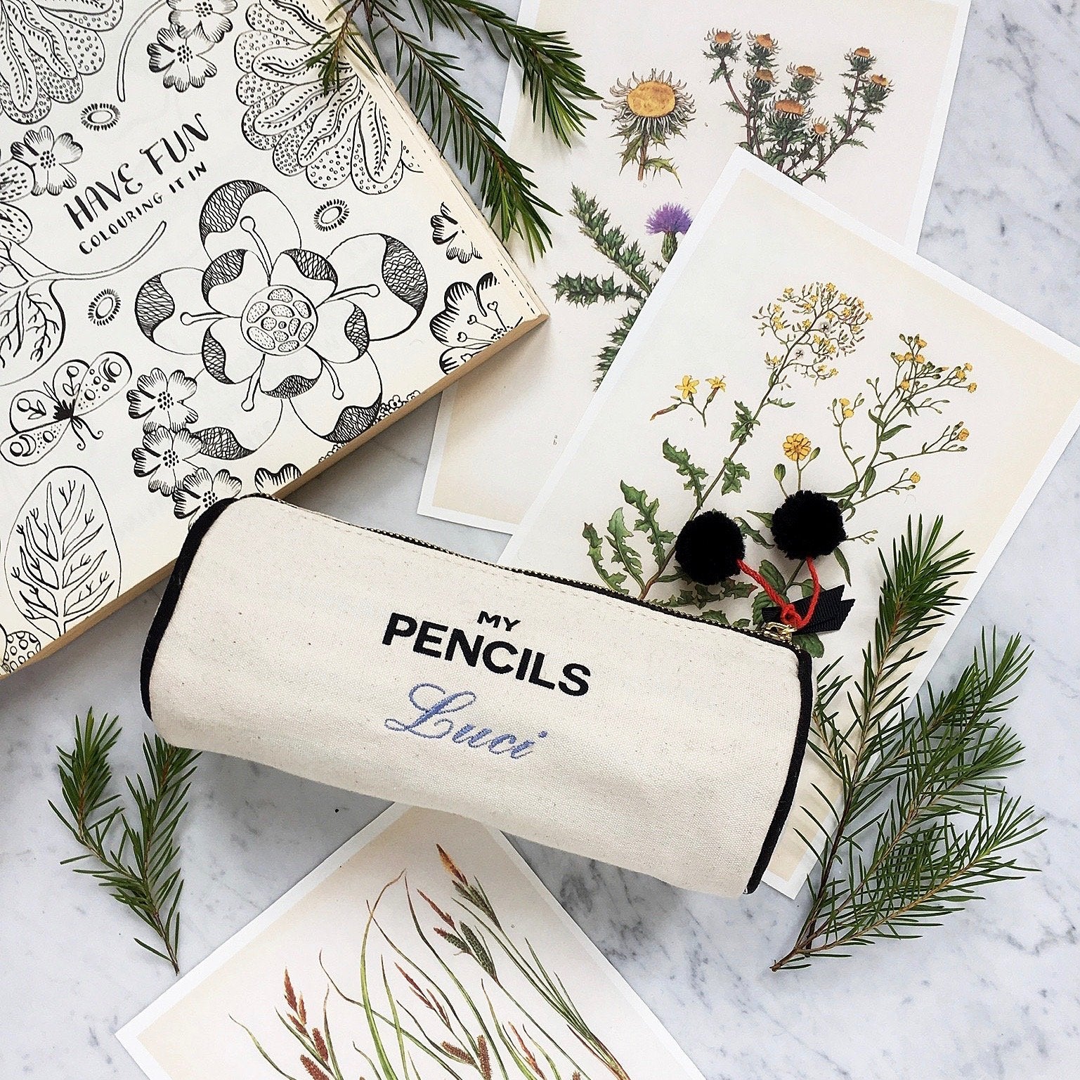 Bag-all cream canvas pencil case with black text reading My Pencils, decorated with pom-poms, displayed on marble surface with botanical illustrations and pine sprigs