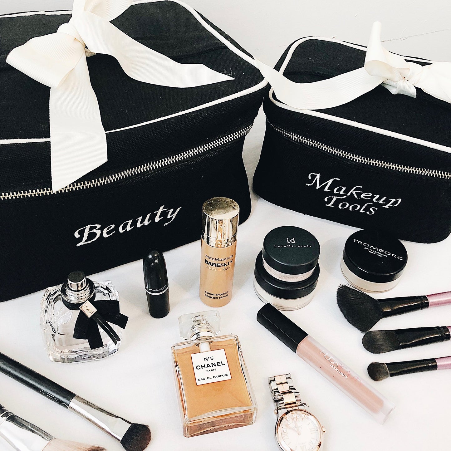Bag-all beauty organizer cases in black with white bow, shown with makeup tools, perfume bottles and cosmetics. Monogram customization available for personalized storage solutions.
