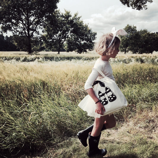 Bag-all Children Shoe Bag in cream color featured in natural outdoor setting, showcasing its practical design with shoe print pattern against golden wheat field backdrop